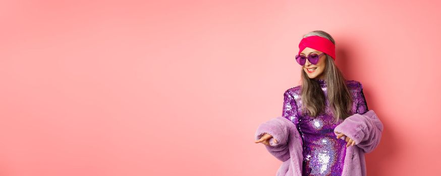 Fashionable asian senior woman dancing disco, wearing glittering shiny dress and heart-shaped sunglasses, having fun, standing over pink background.