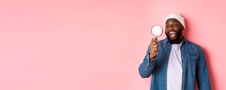 Happy african-american man looking through magnifying glass, smiling amazed, standing against pink background. Copy space
