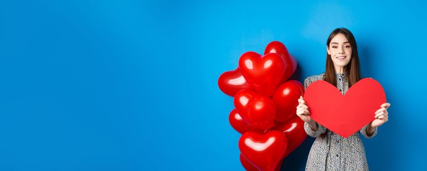 Valentines day. Lovely young woman in dress celebrating lovers holiday, showing valentine card and smiling, standing near red heart balloons on blue background.
