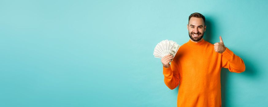 Shopping. Happy man showing thumb up and money in dollars, recommending bank loan or credits, standing over light blue background.