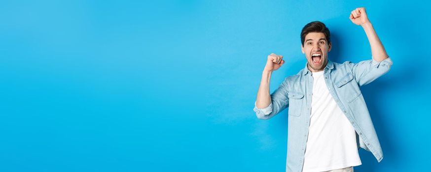 Cheerful guy making fist pumps and rooting for someone, shouting for joy, triumphing over win, standing against blue background.