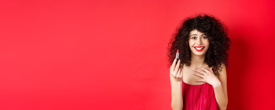 Beauty and make up concept. Beautiful female model with curly hair, wearing evening dress, showing red lipstick and smiling, standing on white background.