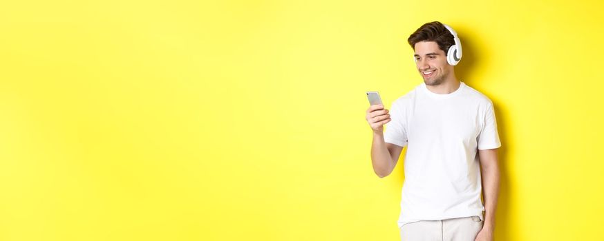 Handsome modern guy choosing playlist on smartphone, wearing headphones, standing over yellow background.