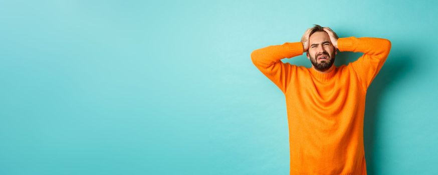 Image of frustrated man in orange sweater, holding hands on head and feeling distressed, standing anxious against turquoise background.