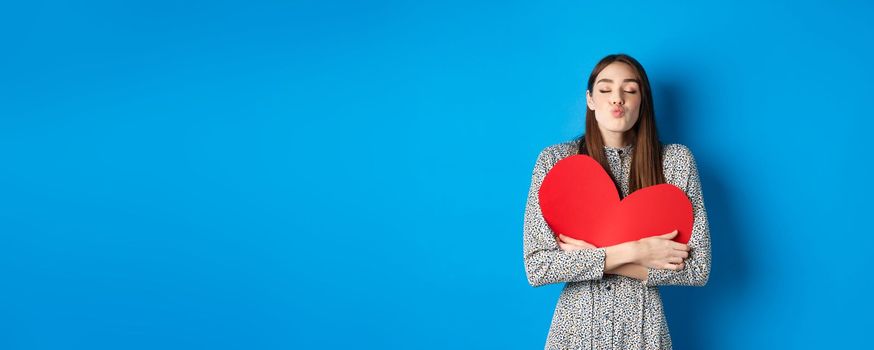 Valentines day. Romantic beautiful woman close eyes and pucker lips for kiss, holding big red heart cutout, kissing you, standing on blue background.