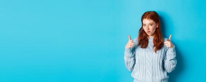 Proud redhead girl pointing fingers at camera, praising and saying congrats, standing over blue background. Copy space