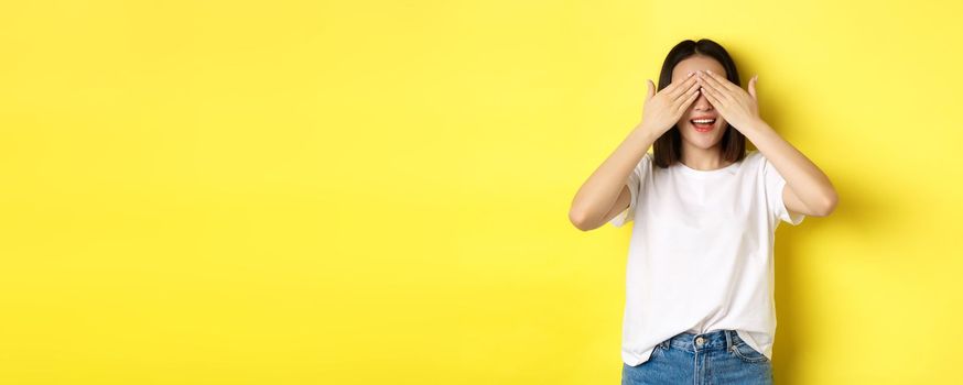 Happy asian woman waiting for surprise gift, close eyes and smiling, expecting something, standing over yellow background.