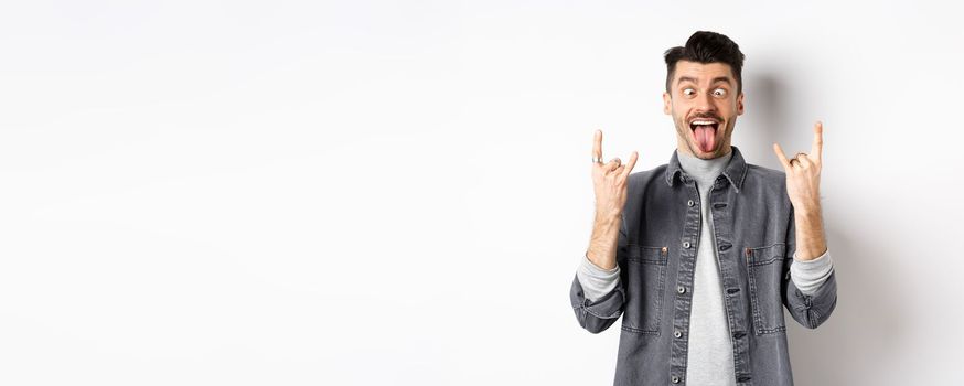 Funny young man showing tongue and rock horns, squinting eyes to fool around, enjoy party or concert, standing on white background.