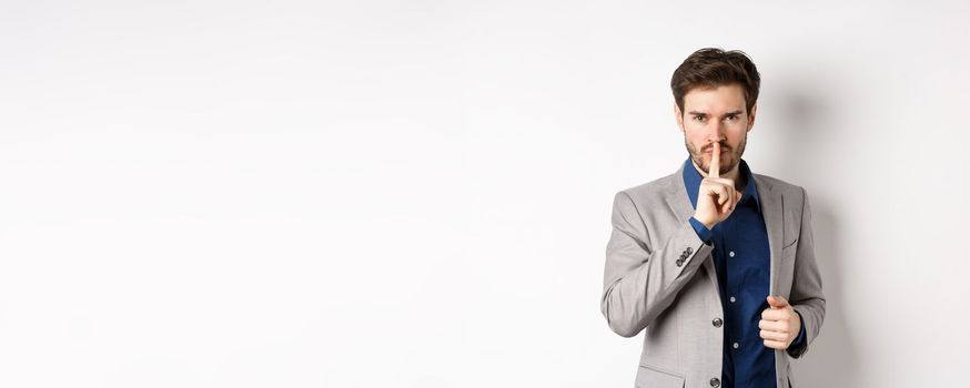 Angry businessman showing taboo sign, shushing and tell to keep quiet, need silence, standing with finger on lips and looking at camera, white background.