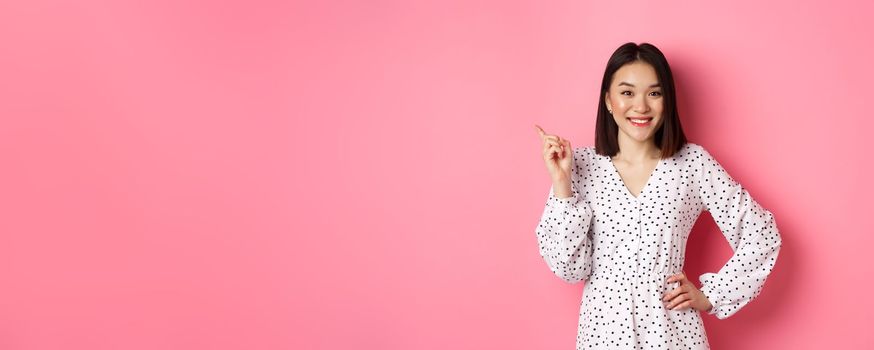Cute asian woman in dress pointing upper left corner copy space. Korean model showing advertisement, standing over pink background.