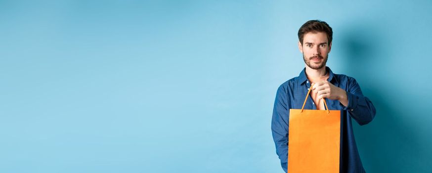 Handsome bearded man stretch out hand with orange shopping bag, making a gift, standing on blue background.