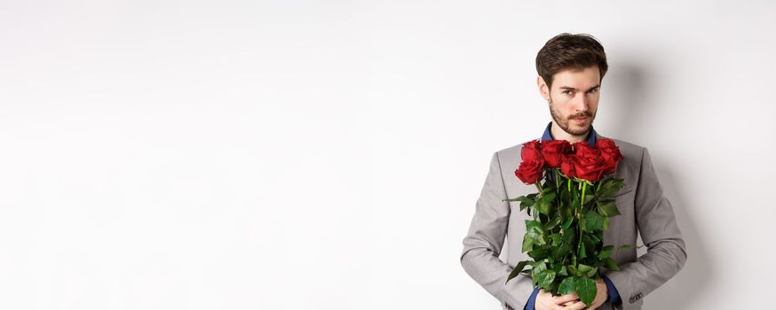 Handsome macho man going on date in suit, holding bouquet of red roses and smiling at camera, making valentines day surprise gift, white background.
