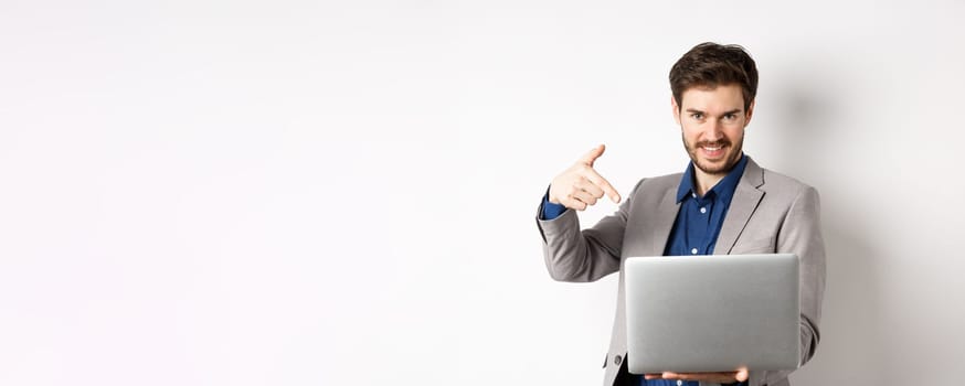 Confident young male entrepreneur working online with laptop, pointing at computer and smiling, standing on white background.