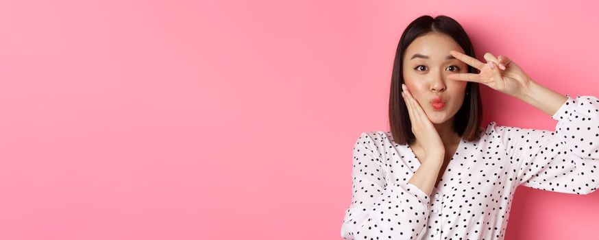 Beauty and lifestyle concept. Close-up of pretty asian woman showing peace sign and touching cheek, smiling happy at camera, standing over pink background.