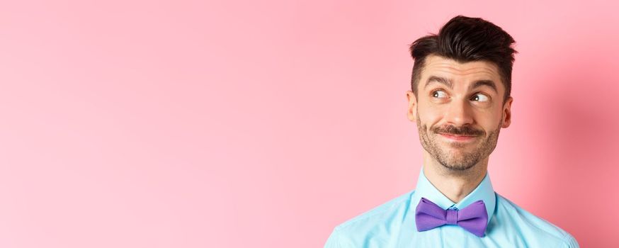 Close-up of handsome smiling guy with moustache, looking left at logo with pleased face, standing on pink background. Copy space