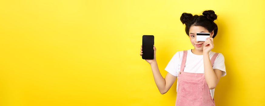 Online shopping concept. Cute asian girl paying with plastic credit card, showing empty smartphone screen and smiling, yellow background.