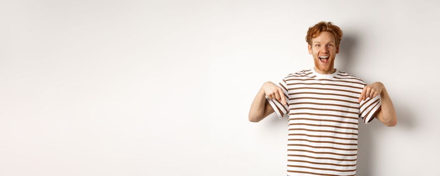 Enthusiastic young man with ginger hair and bristle showing advertisement, pointing fingers down and smiling, white background.