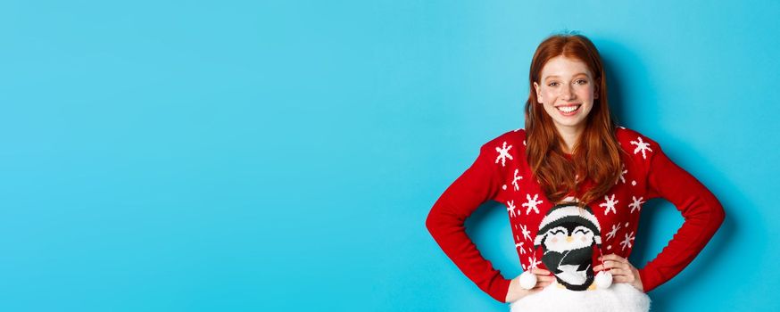 Winter holidays and Christmas Eve concept. Beautiful teenage redhead girl in xmas sweater looking at camera, smiling carefree, holding hands on waist, blue background.