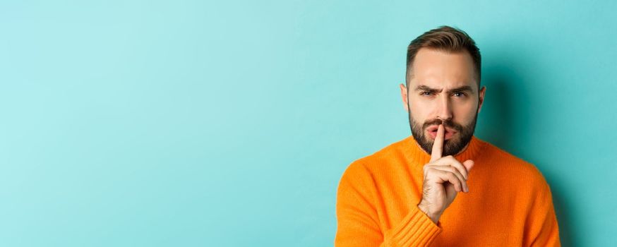 Close-up of annoyed frowning man shushing at camera, press finger to lips, showing taboo gesture, standing over light blue background.