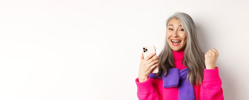 Online shopping. Close-up of cheerful asian elderly woman winning prize in internet, holding smartphone and making fist pump, standing over white background.