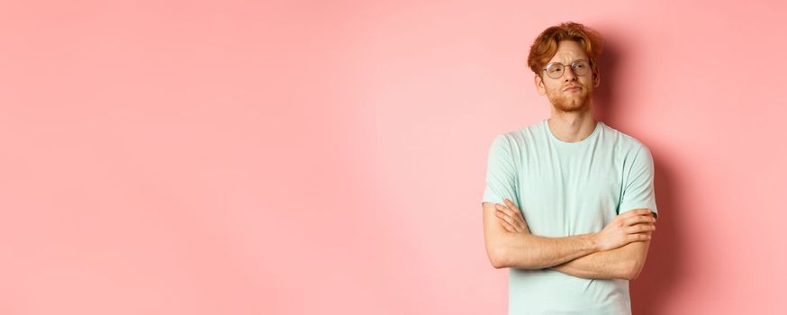 Arrogant redhead guy in glasses cross arms on chest, looking at something with skeptical face, standing over pink background.