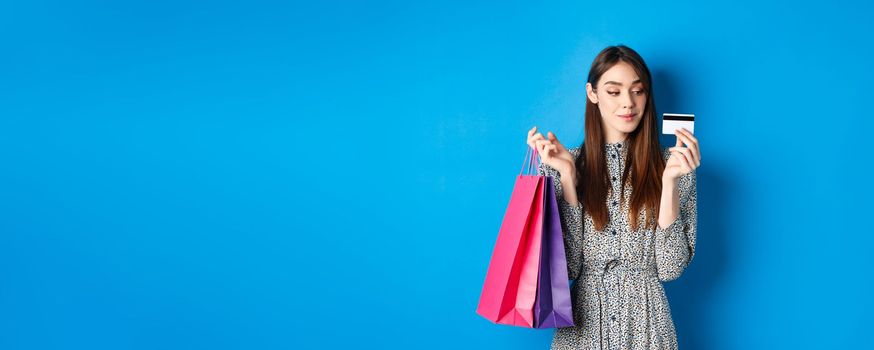 Beautiful girl going on shopping with plastic credit card, holding paper bags with purchased items, standing pleased on blue background.