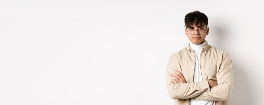 Image of stylish hipster guy in glasses feeling bored, looking unamused with pokerface, cross arms on chest and stand reluctant against white background.