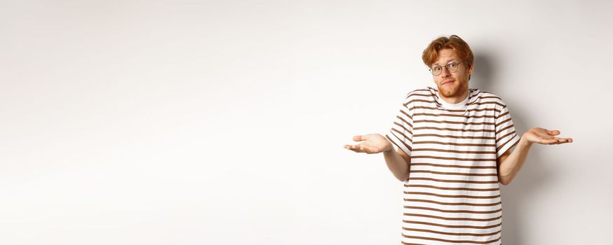 Confused young man with red hair, shrugging shoulders and looking indecisive, standing over white background.