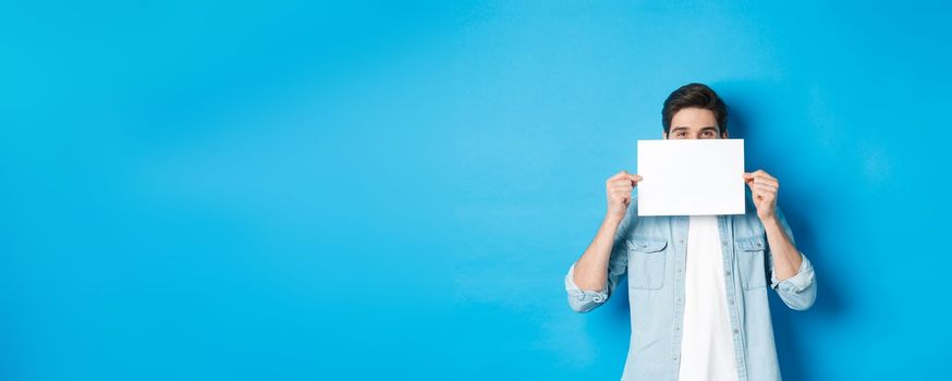 Sneeky handsome guy hiding face behind blank piece of paper for your logo, making announcement or showing promo offer, standing over blue background.