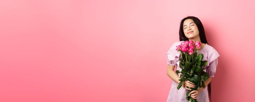Dreamy asian teenage girl feeling romantic, holding flowers and dreaming, imaging valentines day date, wearing cute dress, standing on pink background.