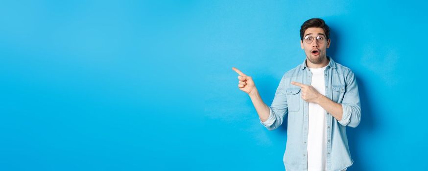 Surprised man in glasses pointing fingers left at copy space, asking question about product, looking intrigued, standing over blue background.