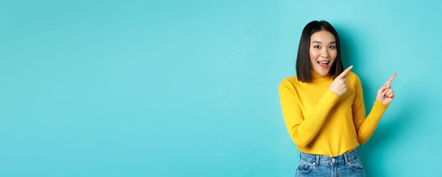 Shopping concept. Beautiful chinese girl in yellow sweater pointing fingers at upper right corner logo banner, smiling amused, standing over blue background.