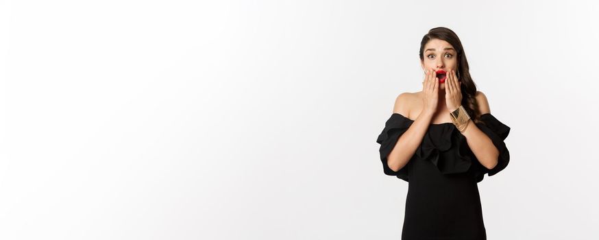 Fashion and beauty. Image of attractive female model in black dress reacting to announcement, looking amazed at camera, standing surprised over white background.