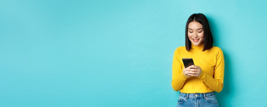 Attractive asian woman reading smartphone screen and smiling, social networking with mobile phone, standing over blue background.