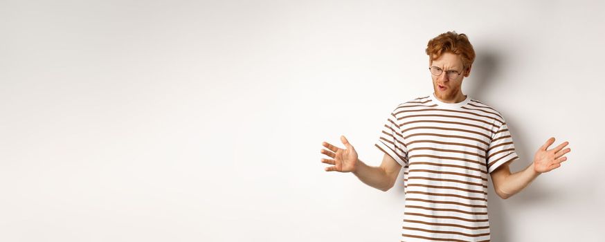 Impressed redhead man in glasses showing length of something big, demonstrate large size and looking down, standing over white background.