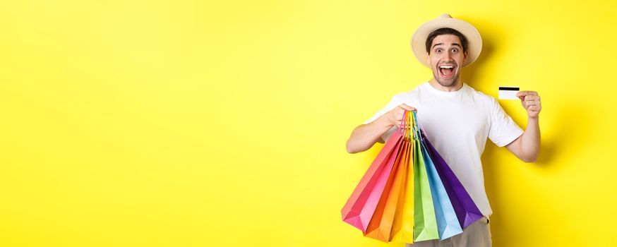 Happy guy going shopping on vacation, holding paper bags and showing credit card, standing over yellow background.