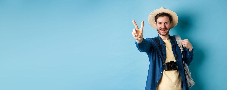 Handsome positive guy tourist showing peace sign on summer vacation, enjoying tropical holidays under sun, wearing straw hat, holding backpack and camera, blue background.