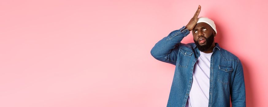 Annoyed Black man roll his eyes and facepalm, standing bothered and tired against pink background.