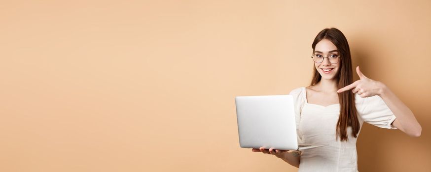 Smiling woman in glasses pointing finger at laptop screen, showing online promo, standing on beige background.