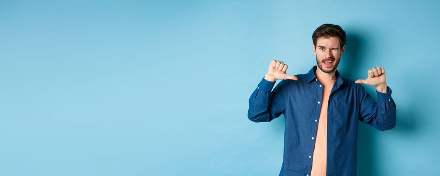 Confident man winking and pointing at himself, self-promoting, standing on blue background. Copy space