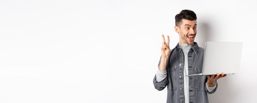 Funny young man video chat on laptop, showing v-sign and smiling at computer camera, standing against white background.