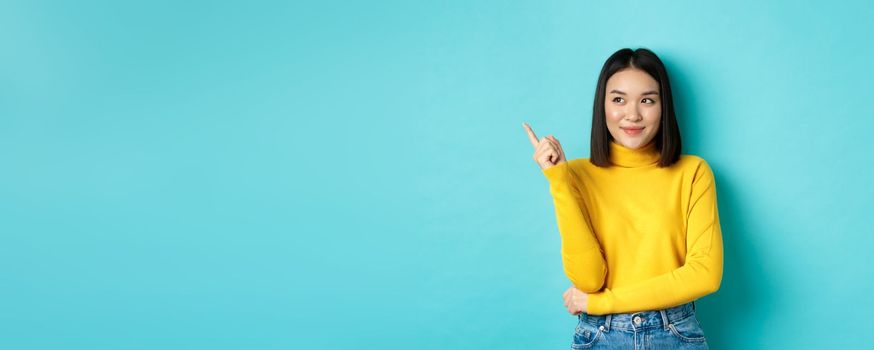 Shopping concept. Stylish asian female model in yellow sweater, smiling and pointing finger left, showing advertisement with satisfied face, standing over blue background.