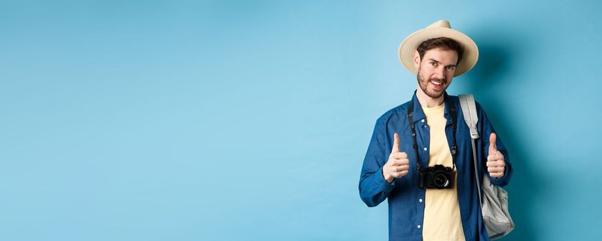 Handsome caucasian tourist in straw hat, holding backpack and photo camera, showing thumbs up, recommending travel agency or hotel resort, blue background.
