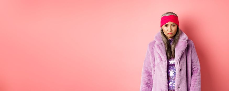 Sad and gloomy stylish asian senior woman looking upset at camera, sulking and feeling distressed, standing in purple faux fur coat against pink background.