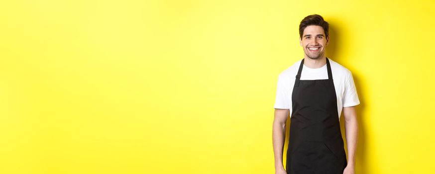 Young man waiter in black apron, smiling, working in store or coffee shop, standing against yellow background.