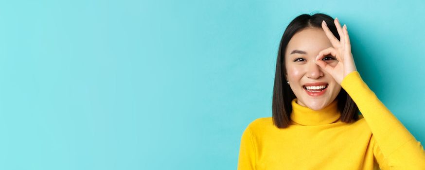 Beauty and makeup concept. Close up of carefree asian girl showing OK sign on eye and smiling, looking happy at camera, standing over blue background.