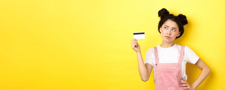 Shopping. Thoughtful asian woman with glamour makeup, holding plastic credit card and looking aside pensive, make decision, yellow background.