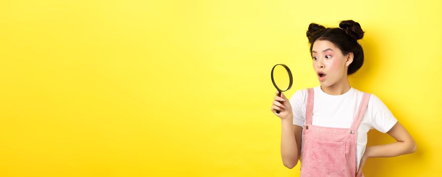 Excited asian woman with bright glamour makeup, look through magnifying glass amazed, staring aside, standing on yellow background.