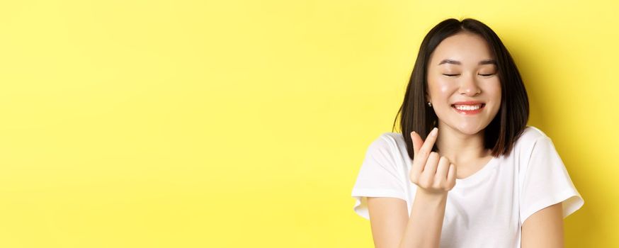Valentines day and women concept. Close up of pretty asian girl in white t-shirt, smiling and showing finger heart, I love you gesture, standing over yellow background.