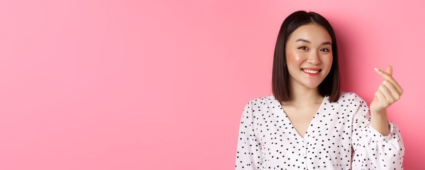 Beauty and lifestyle concept. Close-up of lovely asian woman showing heart sign, smiling and feeling romantic on valentines day, standing over pink background.